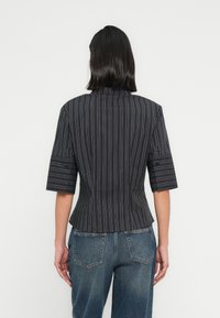 Γυναίκα με σκούρα μαλλιά μέχρι τους ώμους, ντυμένη με μαύρο μπλουζάκι με λεπτές ρίγες και κοντά μανίκια, και μπλε τζιν, γυρισμένη πλάτη σε λευκό φόντο.