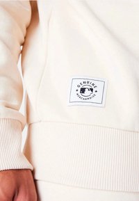 Sudadera de color beige claro con puños y dobladillo acanalados, con una etiqueta cuadrada que muestra "MERCHANDISE GENUINO" y un emblema en negro.