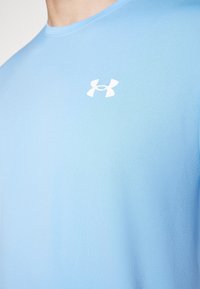 Ljusblå idrottströja i slätt tyg, med en vit logotyp på vänster bröstområde och en klassisk rund halsringning.