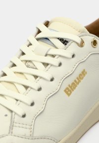 Λευκό δερμάτινο αθλητικό παπούτσι με υφή επιφάνειας, στρογγυλεμένη μύτη και χρυσές λεπτομέρειες. Διαθέτει λευκά κορδόνια και μπλε εσωτερική επένδυση.