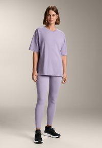 T-shirt in cotone viola, taglio oversize, maniche corte. Abbinato a leggings viola coordinati, texture liscia, e scarpe da ginnastica nere.