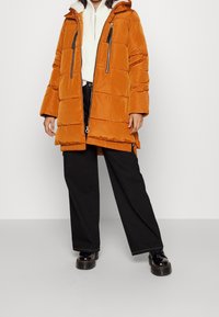 Oranssi puhvijakku, jossa on huppu, vetoketjullinen etuosa ja taskut, yhdistettynä mustiin leveisiin housuihin ja paksuihin mustiin kengät. Rakeinen kangas.