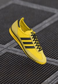 Gule treningssko med svarte detaljer, med tre svarte striper, teksturert overmateriale og lavprofil-design.