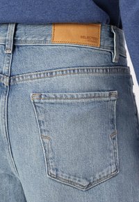Jean en denim bleu clair avec un patch en cuir beige portant l'inscription "SELECTED HOMME", doté de poches arrière classiques et de détails de couture discrets.