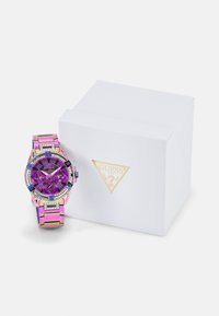 Montre Guess colorée avec un bracelet en acier inoxydable rose et violet et des accents scintillants, présentée aux côtés d'une boîte blanche arborant un logo doré.
