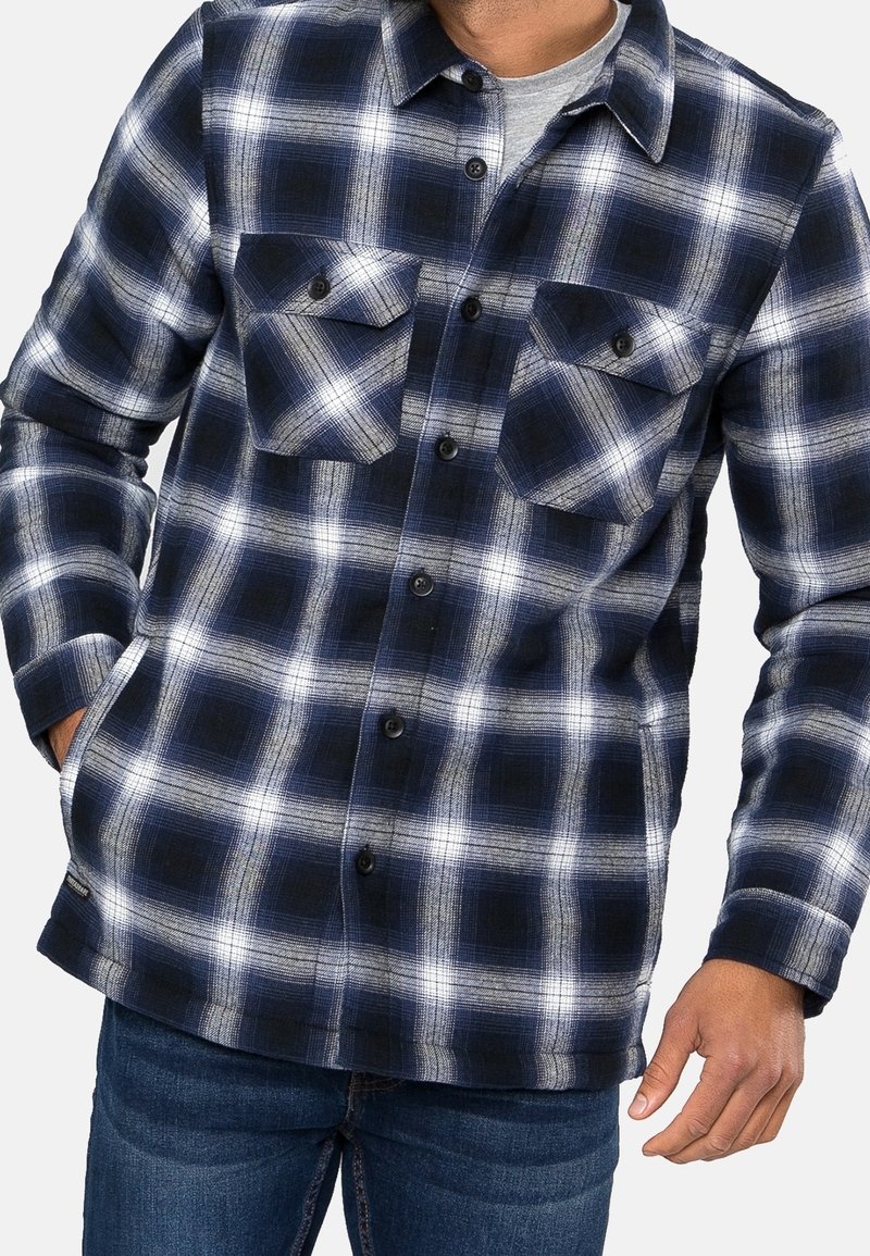 Chemise en flanelle à carreaux bleu marine et blanc avec fermeture boutonnée, deux poches poitrine et manches longues, présentant un ourlet droit et une texture de poids moyen.