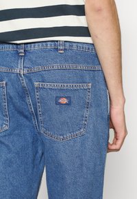 Vue de dos d'une personne portant un jean en denim bleu avec un patch logo Dickies sur la poche arrière droite et une chemise rayée blanche et noire.