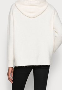 Pull en maille côtelée blanc avec capuche et épaules tombantes ; présente un ourlet légèrement court et un tissu texturé. Porté avec des leggings noirs.