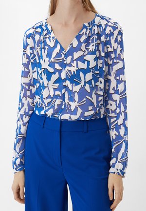 Femme portant un chemisier bleu et blanc à motifs à manches longues, rentré dans un pantalon bleu roi uni, debout devant un fond uni.