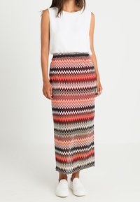 Langer Rock mit Zickzackmuster in Rot, Schwarz und Creme, mit elastischem Bund, kombiniert mit einem ärmellosen weißen Oberteil und weißen Sneakers.