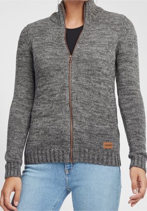 Femme portant un pull gris en tricot zippé avec une étiquette en cuir de la marque et un jean bleu clair, debout devant un fond uni.