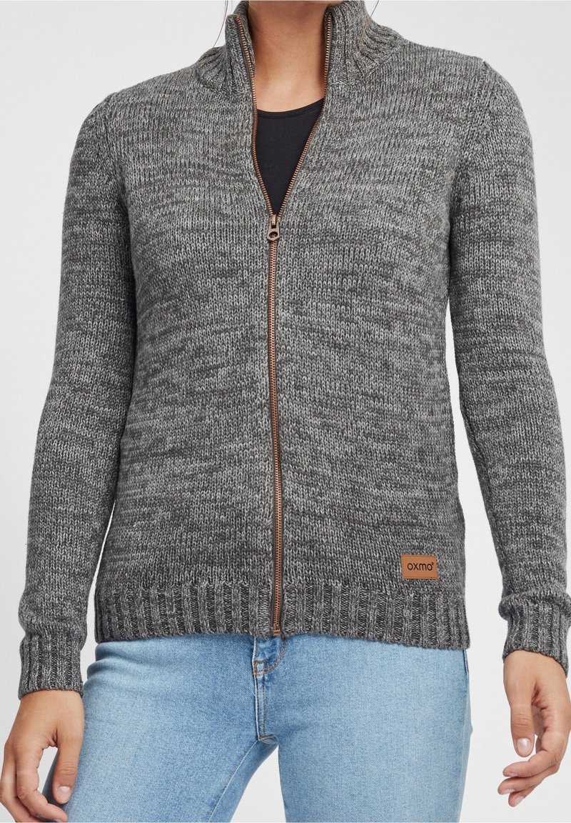 Femme portant un pull gris en tricot zippé avec une étiquette en cuir de la marque et un jean bleu clair, debout devant un fond uni.