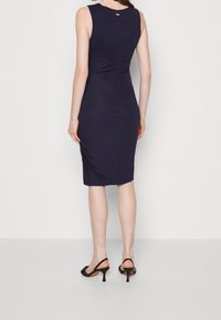 Kvinna iklädd en åtsittande ärmlös marinblå klänning och svarta klacksandaler, stående mot en enfärgad ljus bakgrund, med ryggen vänd mot kameran.