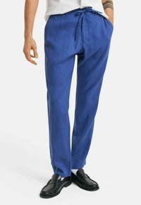 Blauwe linnen broek met een trekkoord in de taille, voorzien van twee zijzakken en een losse pasvorm. Draagt een paar zwarte instappers op een witte achtergrond.