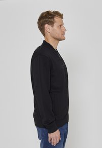 Blouson aviateur noir en tissu lisse avec fermeture éclair et poches latérales. Doté d'un col et de poignets côtelés, porté avec un jean bleu.