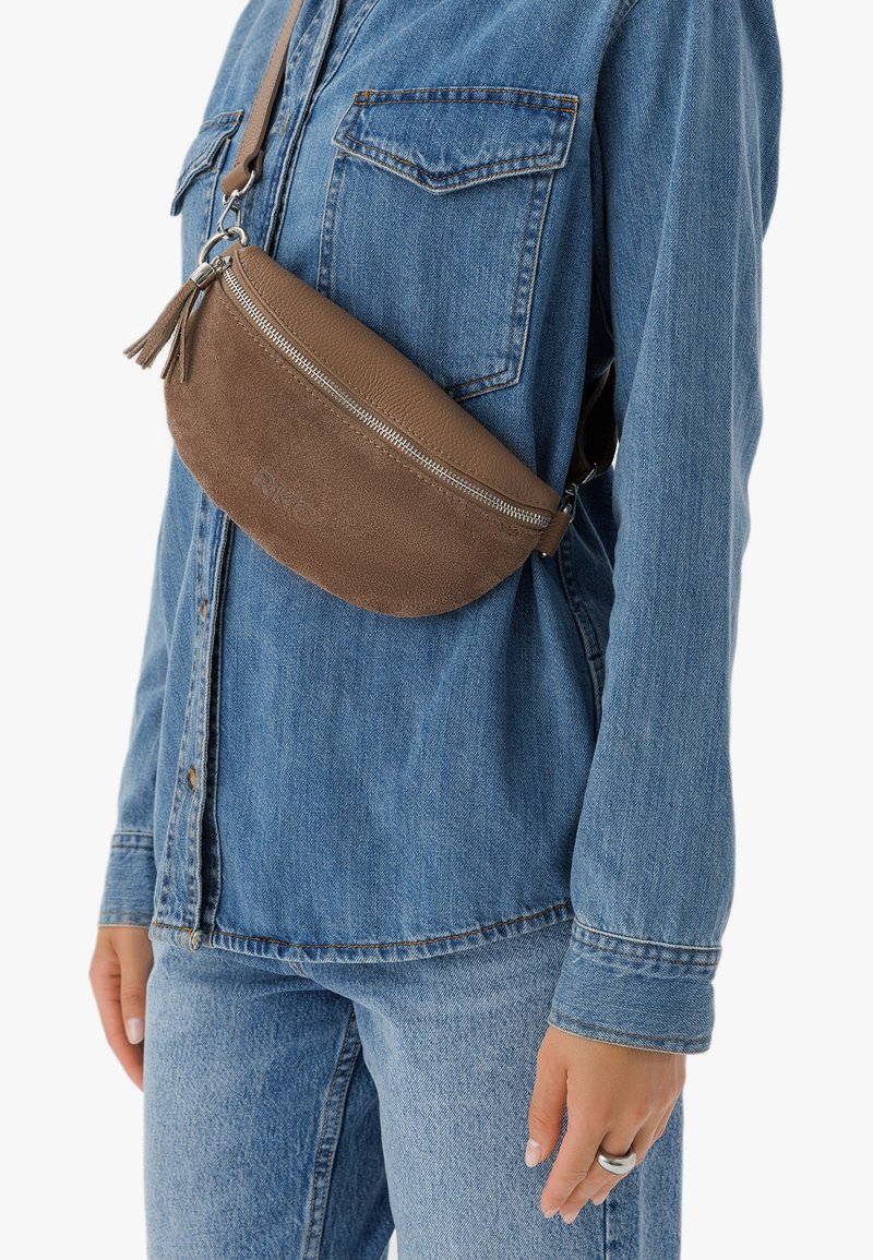 Ruskea nahkainen vyölaukku, jossa on vetoketju ja tupsuaksentti, käytetty denim-paitojen ja farkkujen päällä, esittäen sileää pintaa ja pyöreää muotoa.