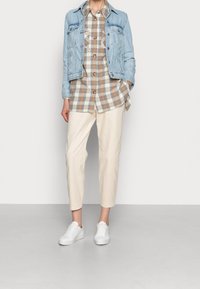 Persoon in een lichtblauwe spijkerjack, met een bruin-witte geblokte shirt, beige broek en witte sneakers, staand tegen een effen witte achtergrond.