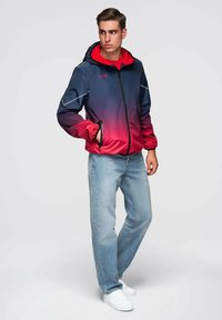 Mladý muž v kapucovém blejzru s modro-červeným ombré, světlých modrých džínách a bílých teniskách, stojící s jednou rukou v kapse.