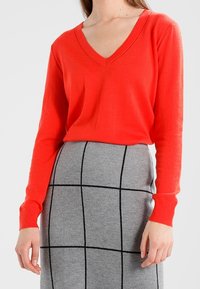 Femme portant un pull rouge vif à col en V et manches longues, ainsi qu'une jupe grise taille haute avec un motif à carreaux noirs.