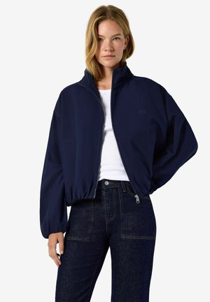 Femme aux cheveux blonds mi-longs portant une veste zippée bleu marine, un haut blanc et un jean taille haute bleu foncé sur un fond uni.