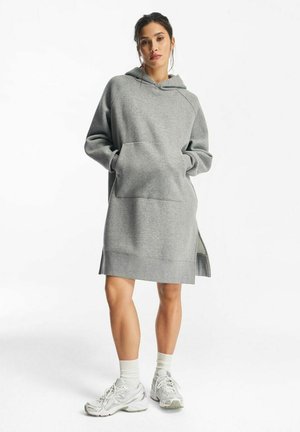 Kobieta ubrana w szarą sukienkę z kapturem i kieszenią z przodu, białe skarpetki i białe trampki, stojąca na jednolitym białym tle.