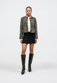 Chaqueta corta con estampado de cebra en negro y beige, top blanco, falda negra plisada y botas negras hasta la rodilla con puntera puntiaguda.