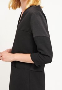 Robe noire avec décolleté en V, manches trois-quarts et design structuré. Présente des panneaux texturés contrastants dans la partie supérieure.