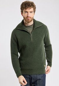 Pull à col haut en tricot côtelé vert, avec une fermeture éclair à mi-hauteur à l'avant et une petite étiquette logo au bas. Porté par-dessus une chemise verte assortie.