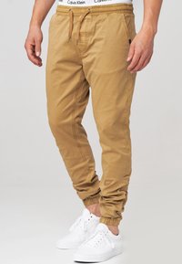Pantalones jogger en color caqui con puños elásticos y cintura con cordón, que presentan una suave tela de algodón y bolsillos laterales, combinados con zapatillas deportivas blancas.