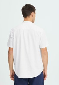 Chemise blanche en lin à manches courtes avec empiècement arrière et ourlet arrondi. Le tissu présente une apparence légère et texturée.