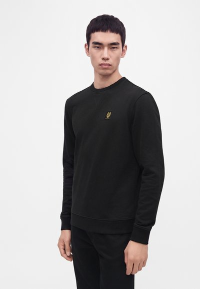 Jeune homme aux cheveux noirs courts portant un sweat-shirt noir à manches longues avec un petit emblème doré sur la poitrine, debout devant un fond blanc uni.