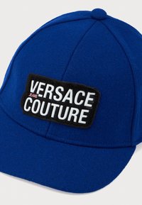 Modrá plstená baseballová čiapka s čiernym nášivkom a bielym textom "VERSACE JEANS COUTURE." Zaoblený šilt a prešité detaily.