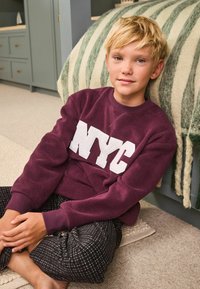 Sweatshirt bordeaux avec lettrage blanc "NYC", associé à un pantalon à carreaux noir et blanc. Tissu doux et texturé, coupe décontractée.