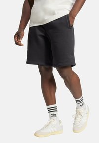 Shorts en coton noirs avec poches latérales, présentant un détail de logo texturé. Associés à des chaussettes rayées et des baskets blanches avec des accents jaunes.