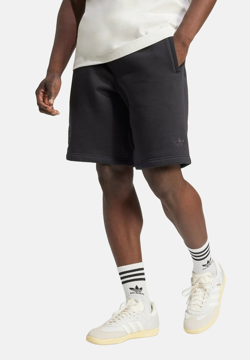 Shorts en coton noirs avec poches latérales, présentant un détail de logo texturé. Associés à des chaussettes rayées et des baskets blanches avec des accents jaunes.