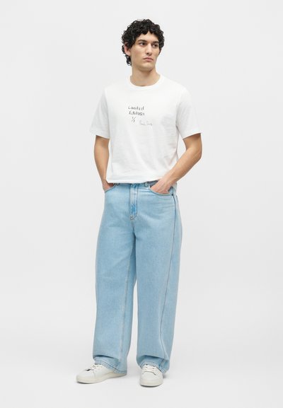 Άνδρας με σγουρά μαλλιά που φοράει λευκό μπλουζάκι "Limited Edition", φαρδιά ανοιχτό μπλε τζιν και λευκά αθλητικά, στέκεται με τα χέρια στις τσέπες.