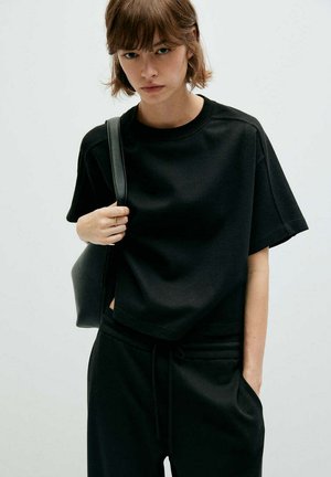 Jeune femme en chemise noire à manches courtes et pantalon porte un sac à bandoulière noir avec une main dans la poche devant un fond uni.