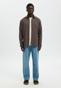 Brun skjorta med knappar och långa ärmar, ljusbeige t-shirt under, ljusblå vida jeans och svarta sandaler med strukturerad sula.