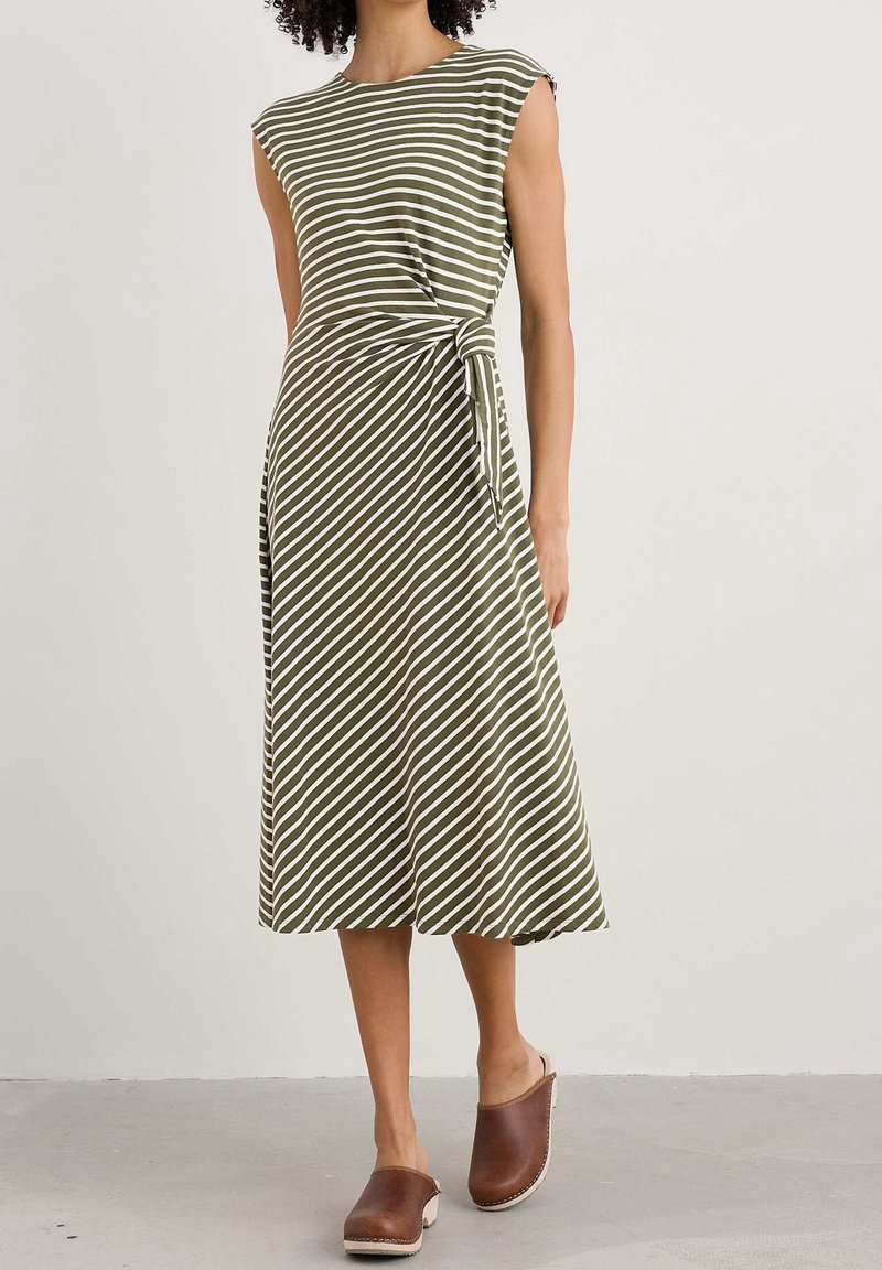 Femme portant une robe midi sans manches à rayures vert olive et blanc avec un lien sur le côté, associée à des sabots marron à enfiler.