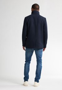 Marineblauer Wollmischungs-Mantel mit hohem Kragen, taillierter Form und Rückennahtdetails, kombiniert mit blauen Jeans und weißen Sneakers.