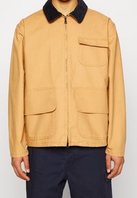 Tan zip-up jacket with a corduroy collar, featuring four front pockets and a durable fabric texture. Suitable for casual wear.
