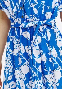Niebieska sukienka z kwiatowym białym wzorem, zapięciem na guziki, podkreśloną talią z dopasowanym paskiem oraz drapowaną, zwiewną sylwetką.