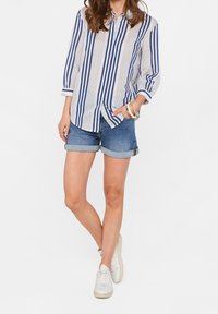 Blau-weiß gestreiftes Hemd mit Knöpfen und hochgekrempelten Ärmeln, kombiniert mit Jeans-Shorts. Das Model trägt wei ße Turnschuhe und Accessoires.