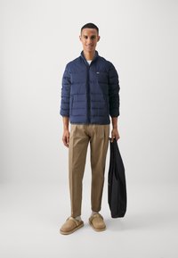 Marineblå pufferjakke med horisontal quilt, lett innerforing, matchende beige bukser og brune slip-on sko, holdt med en svart klespose.