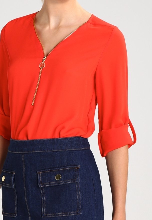 Röd blus med V-hals och dragkedjedetalj. Har trekvartsärmar med uppvikta manschetter. Kombinerad med en mörk denimkjol med fickor.