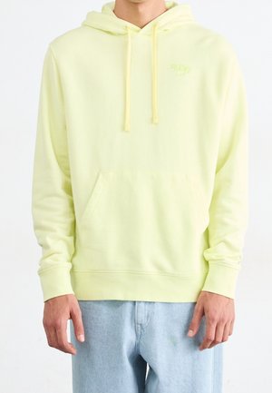 Sweat à capuche jaune clair avec poche avant et cordons porté par une personne en jean bleu clair sur fond blanc.