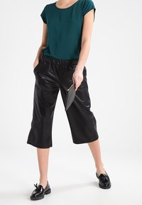 Zwarte cropped broeken met een glanzende textuur, elastische tailleband, gecombineerd met een teal blouse en zwarte schoenen. Het model houdt een kleine clutch vast.