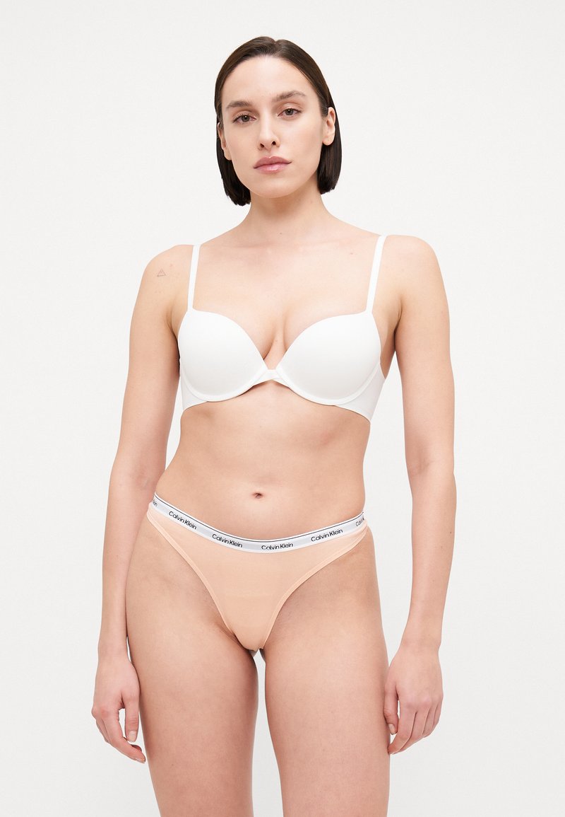 Bela podložena modrčka z nastavljivimi naramnicami, v kombinaciji s golim tangicami z elastičnim pasom s Calvin Klein logotipom. Gladka tekstura, minimalističen dizajn.