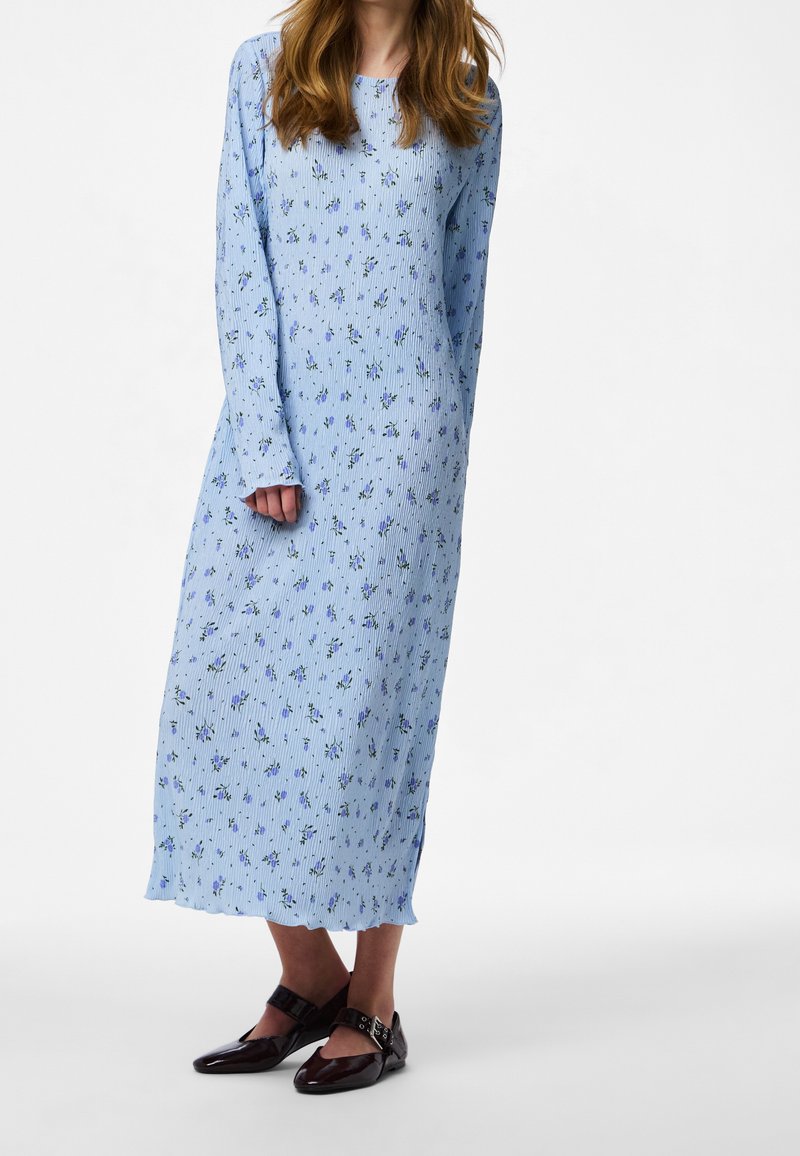 Femme portant une longue robe fleurie bleu clair à manches longues et des chaussures Mary Jane sombres et brillantes, debout devant un fond uni.