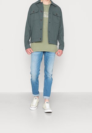 Tumma vihreä napillinen takki, jossa neljä taskua, kerroksittain vihreän paidan päällä; vaaleansininen kapenevat farkut ja valkoiset lenkkarit.